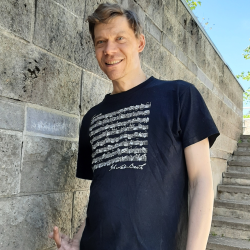 Henrikki, vaalean ruskeat hiukset, tummansininen paita jossa kuvioita, seisoo harmaan kiviseinän edessä, taustalla portaat.