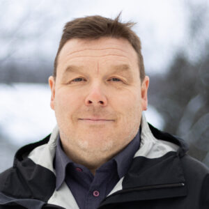 Hymyilevä mies jolla tummat hiukset, kauluspaita ja musta takki. Taustalla luminen maisema.