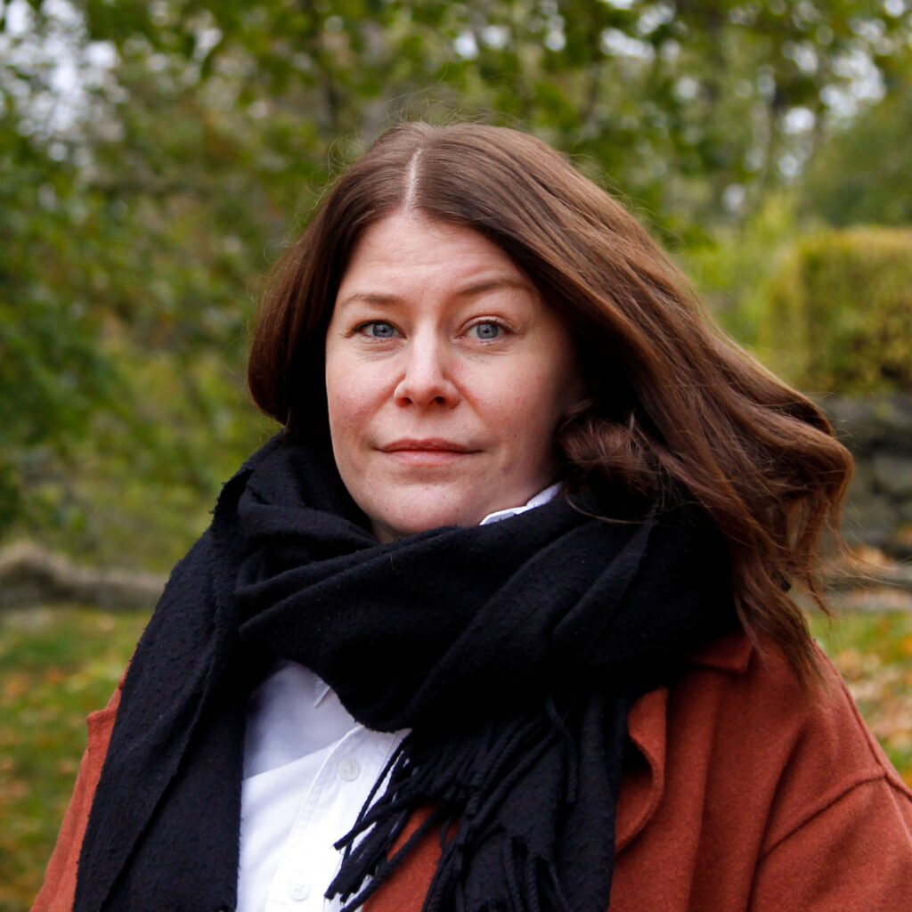 Ruskeahiuksinen nainen, jolla on musta huivi ja punaruskea takki. Taustalla vihreälehtinen puu.