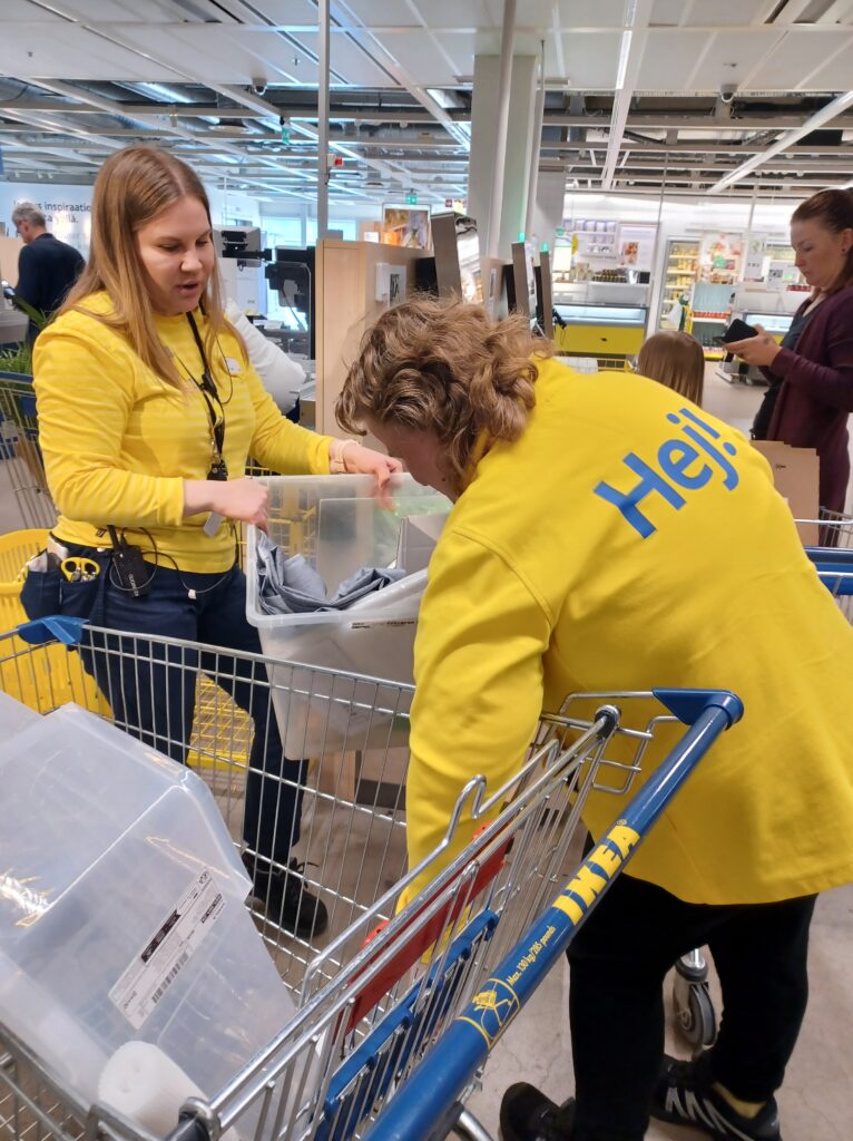 Kaksi naista on asiakkaiden joukossa. Yhdellä on ostoskärryt. Toinen heistä kantaa muovista, läpinäkyvää laatikkoa käsissään.