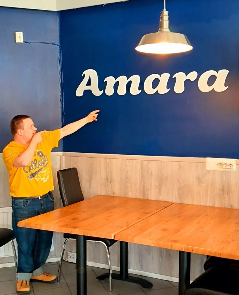 Mies osoittaa seinässä olevaa pizzerian "Amara"-logoa.