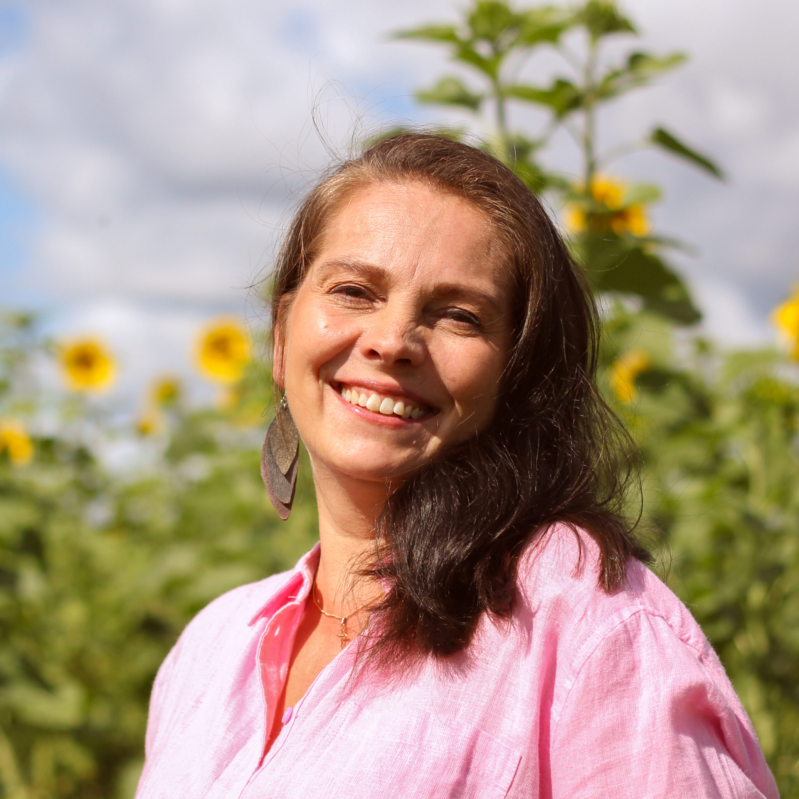 Ruskeat pitkät hiukset, vaaleanpunainen paitapusero, nainen hymyilee auringonkukkia taustalla.