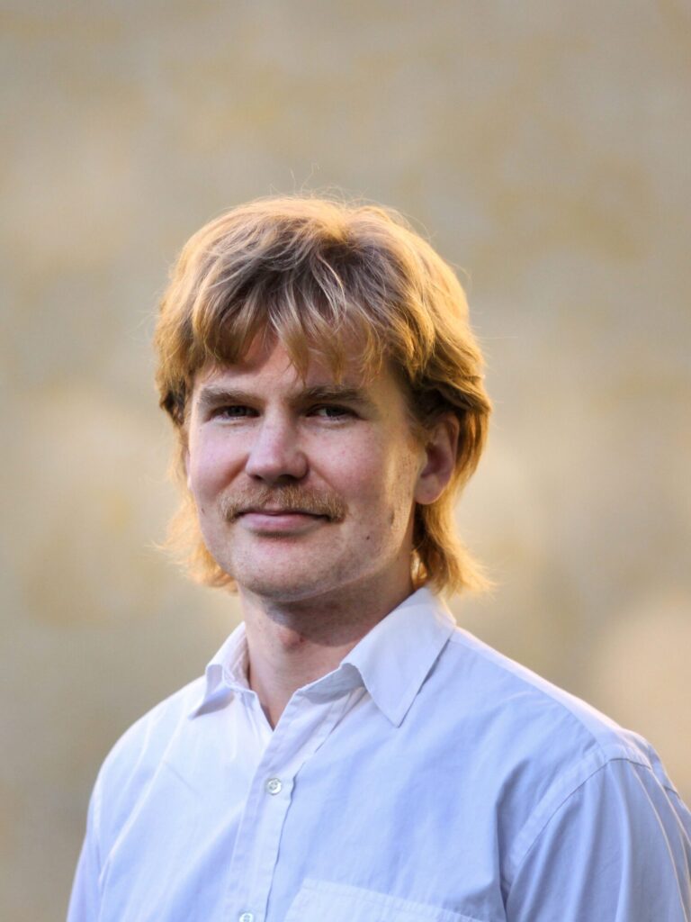 Mies valkoinen kauluspaita päällä. Hymyilee ja katsoo kameraan.