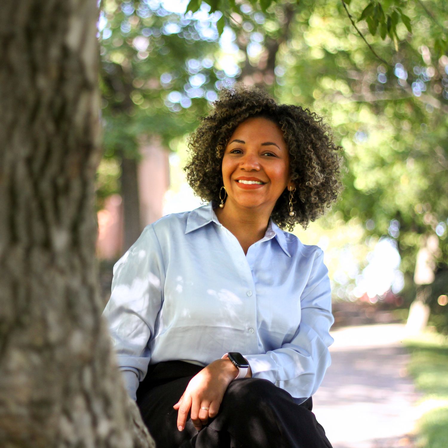 Hymyilevä nainen istuu puunrungolla ja katsoo kameraan. Päällä vaaleansininen kauluspaita.