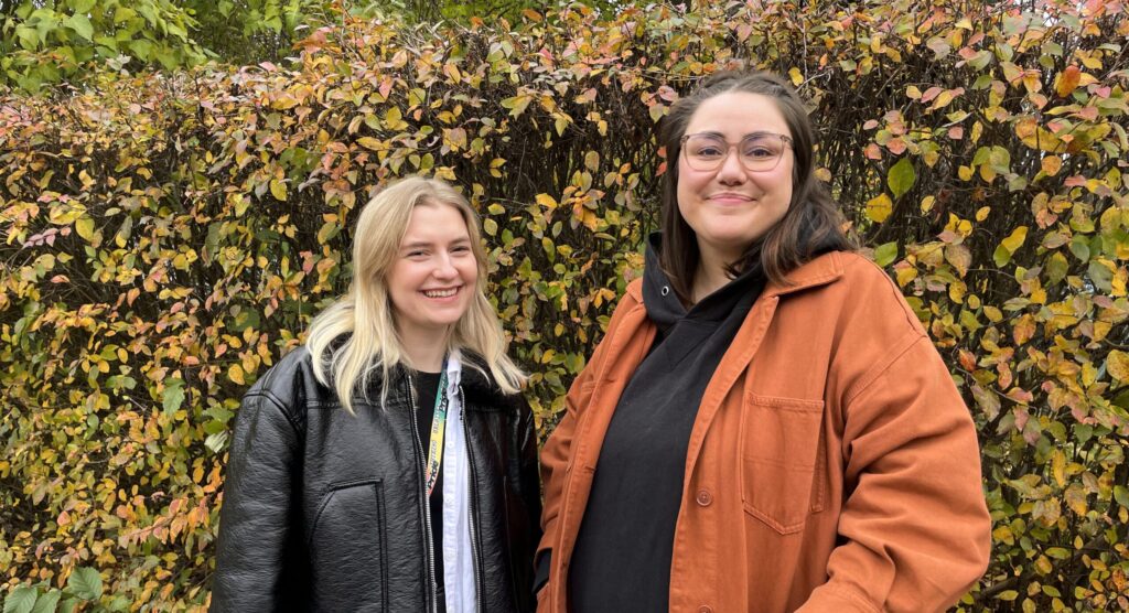 Vasemmalla Johanna ja oikealla Iida. Naiset hymyilevät syksyn ruskan värittämän pensasaidan edessä.