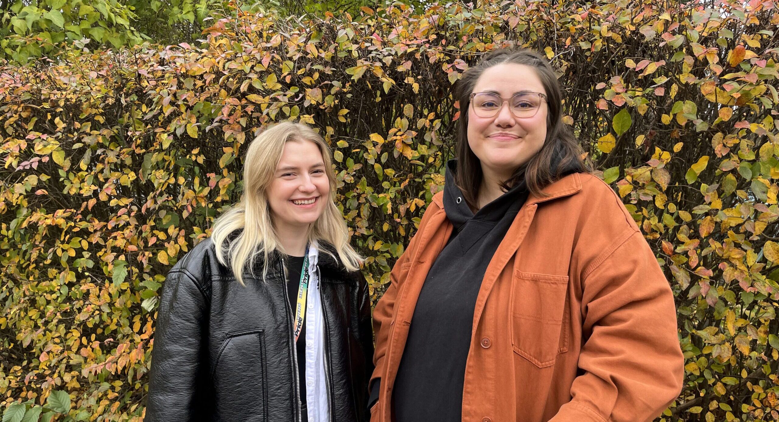 Vasemmalla Johanna ja oikealla Iida. Naiset hymyilevät syksyn ruskan värittämän pensasaidan edessä.