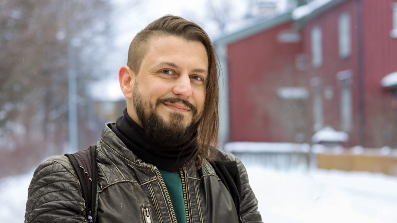 Tummahiuksinen parrakas mies, jolla ruskea nahkatakki.