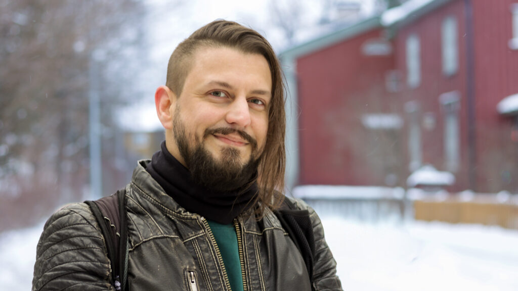 Tummahiuksinen mies, jolla parta, viikset ja ruskea nahkatakki. Taustalla luminen maisema ja punainen talo.