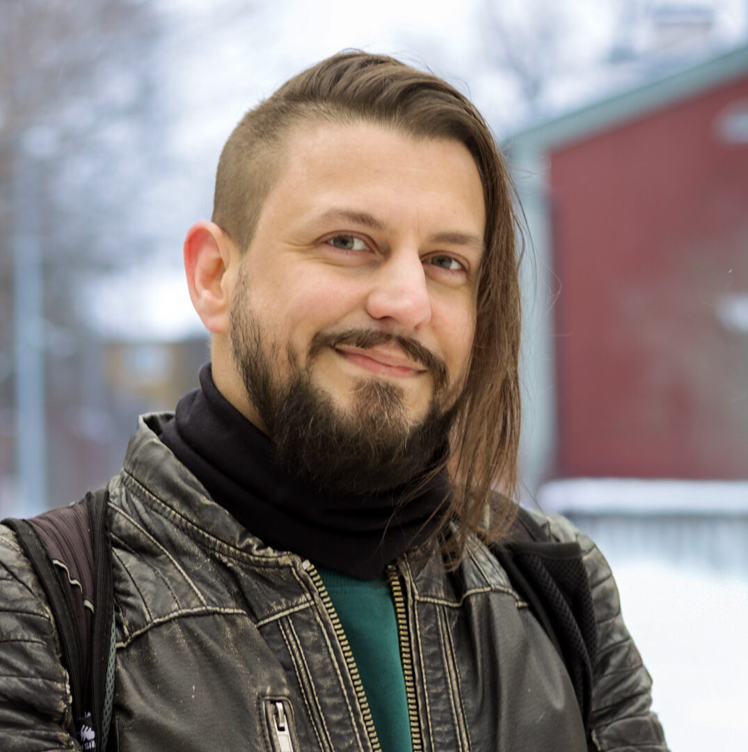 Tummahiuksinen mies, jolla parta, viikset ja musta nahkatakki. Taustalla luminen maisema ja punainen puutalo.