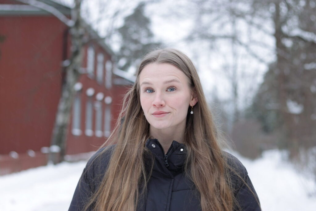 Pitkähiuksinen ja sinisilmäinen nainen hymyilee punaisen talon edustalla.
