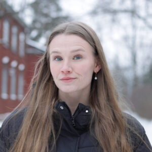 Pitkähiuksinen ja sinisilmäinen nainen hymyilee punaisen talon edustalla.