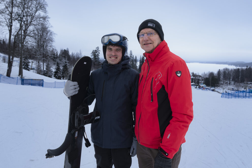 Tummiin lasketteluvaatteisiin pukeutunut mies hymyilee ja pitää kädessään lumilautaa. Oikealla puolella punaiseen takkiin pukeutunut isä hymyilee ja katsoo kameraan.
