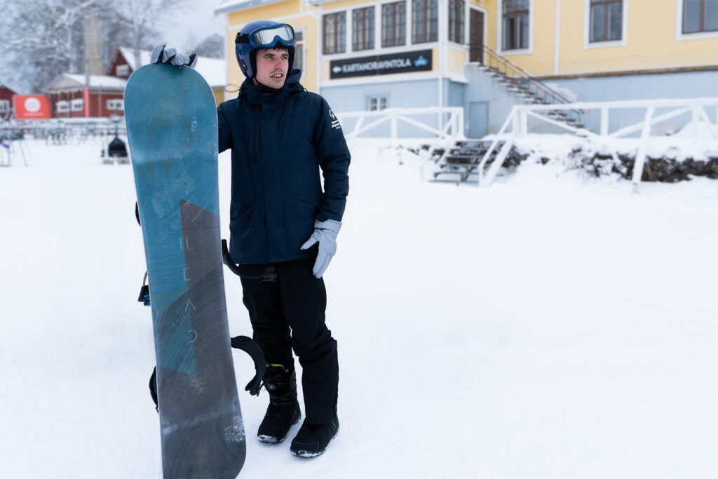 Henkilö seisoo laskettelurinteen päällä ja nojaa oikealla kädellä lumilautaan.