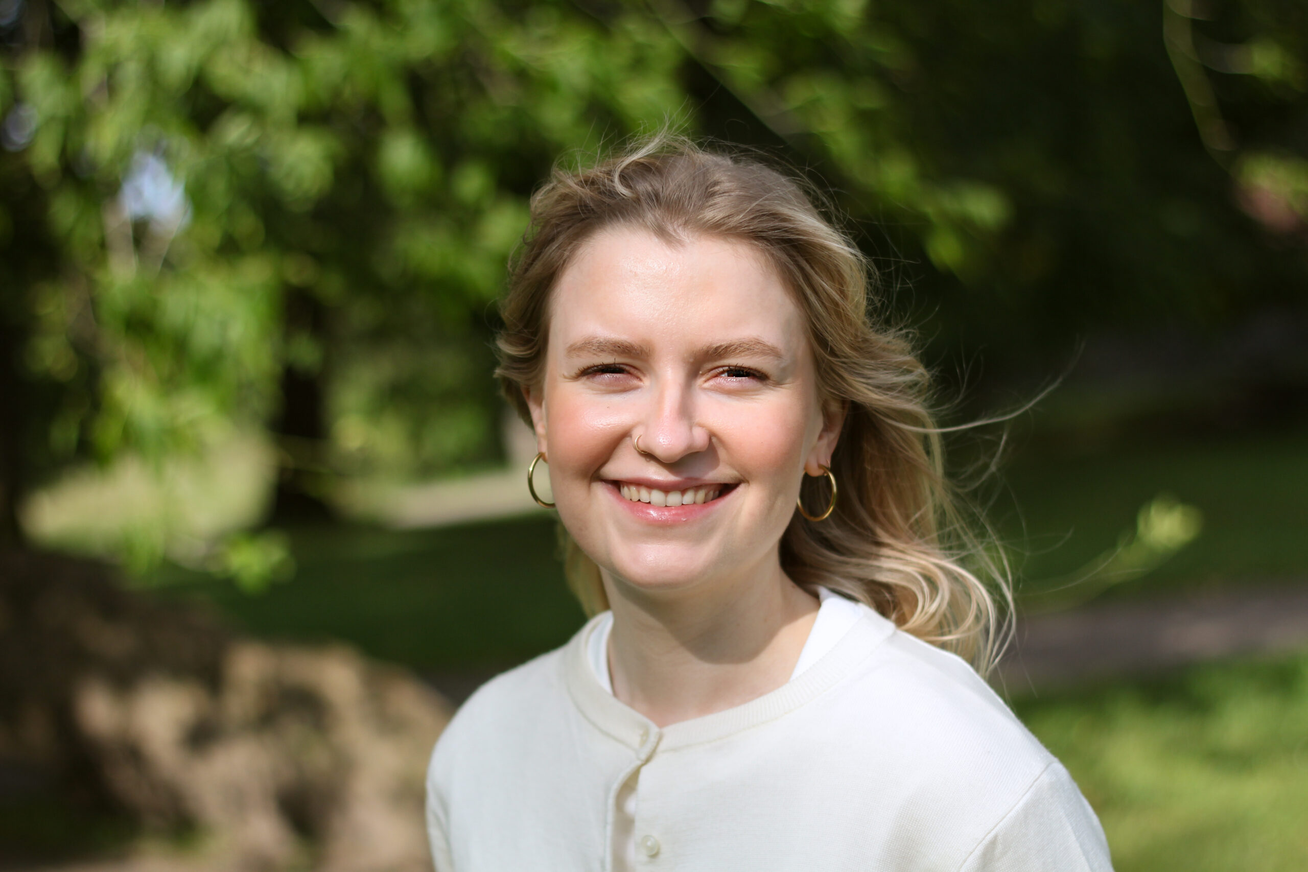 Vaaleahiuksinen ja valkoisessa paidassa nainen puolilähikuvassa hymyilee kesällä vehreässä puistossa, jossa puu taustalla.