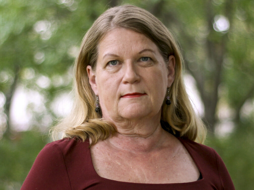 Vaaleahiuksinen nainen viininpunaisessa paidassa kesäisessä puistossa, vihreälehtinen puu taustalla.