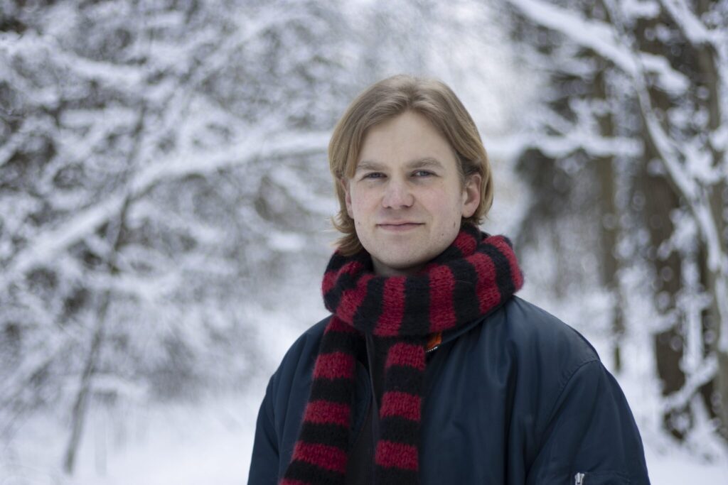Mies hymyilee ja katsoo kameraan. Päällään hänellä on punamusta kaulahuivi ja tummansininen takki. Taustalla lumista metsää.