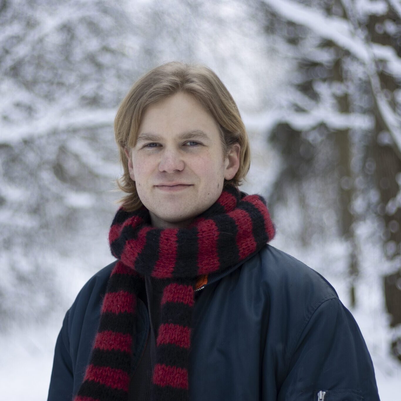 Mies hymyilee ja katsoo kameraan. Päällään hänellä on punamusta kaulahuivi ja tummansininen takki. Taustalla lumista metsää.