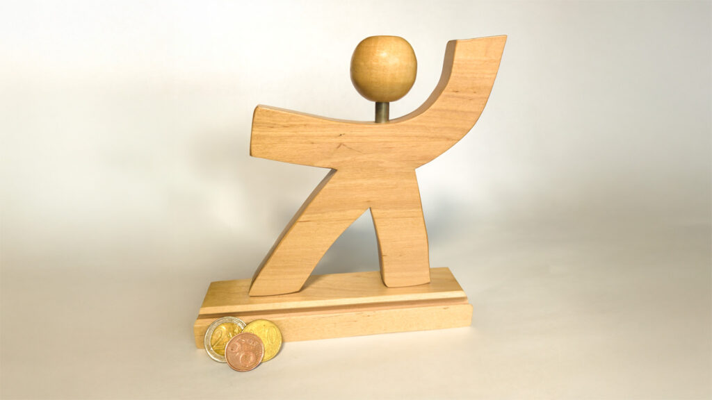 Vatesin puinen logo, jossa hahmon vasen käsi on kohti taivasta. Kuvan vasemmassa reunassa kolikoita.