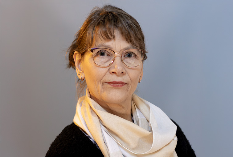 Ruskeahiuksinen nainen, jolla on silmälasit, musta paita ja vaalea huivi katsoo kameraan vakava ilme kasvoillaan.