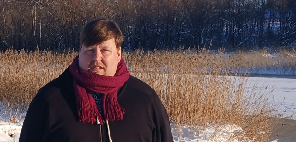 Ilpo Miettinen ulkona joen rannassa. Aurinko paistaa, hanki hohtaa ja taustalla on jäätynyttä kaislaa.