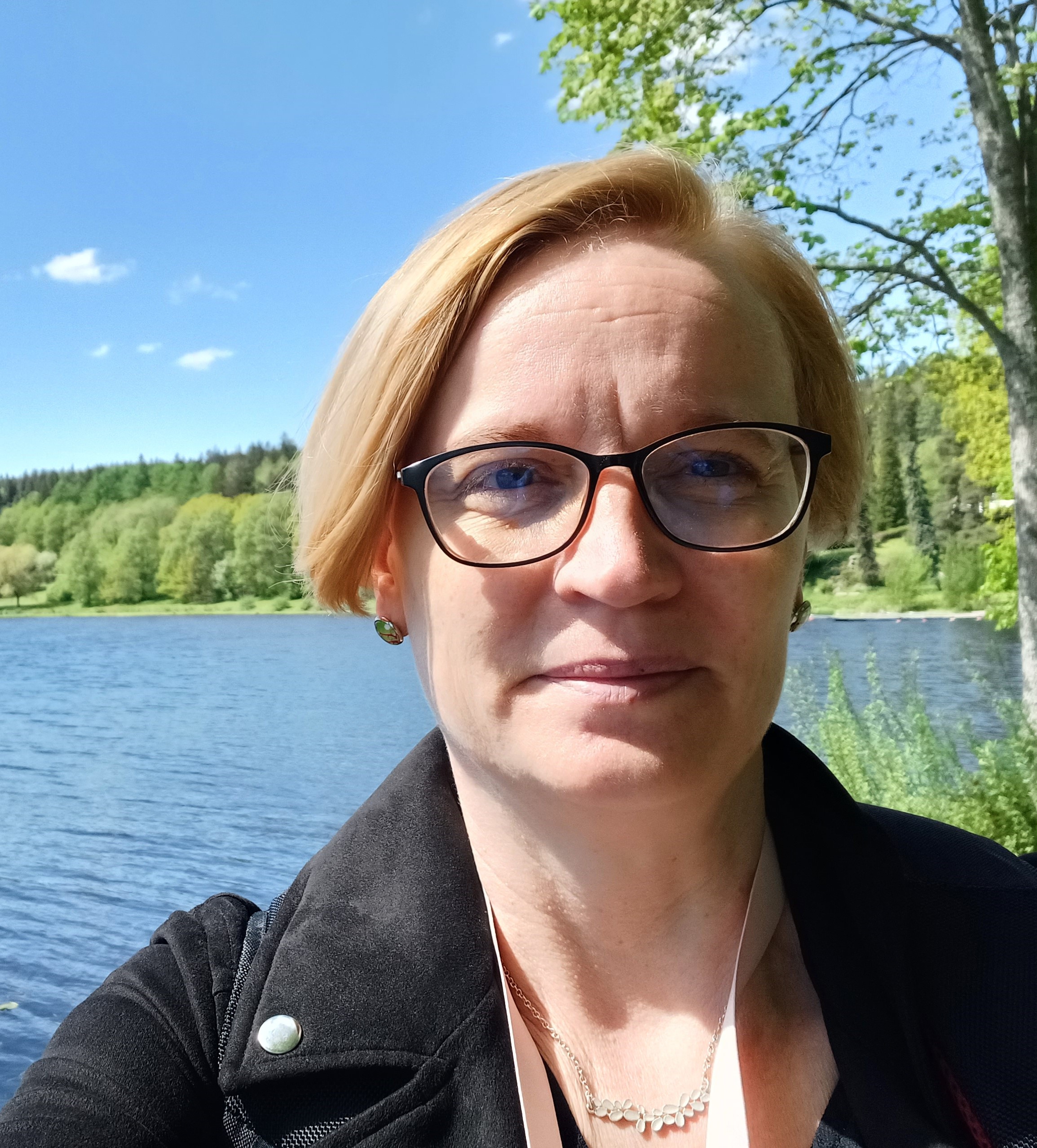 Vaaleahiuksinen ja silmälasipäinen nainen kesäinen järvimaisema taustallaan.