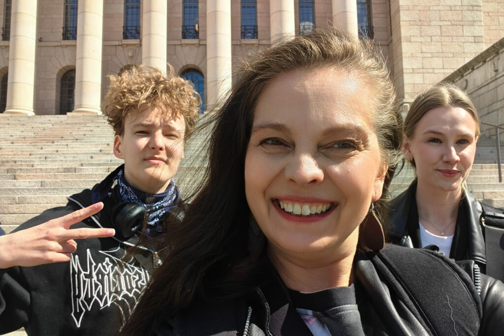 Kolme hymyilevää henkilöä eduskuntatalon edessä.
