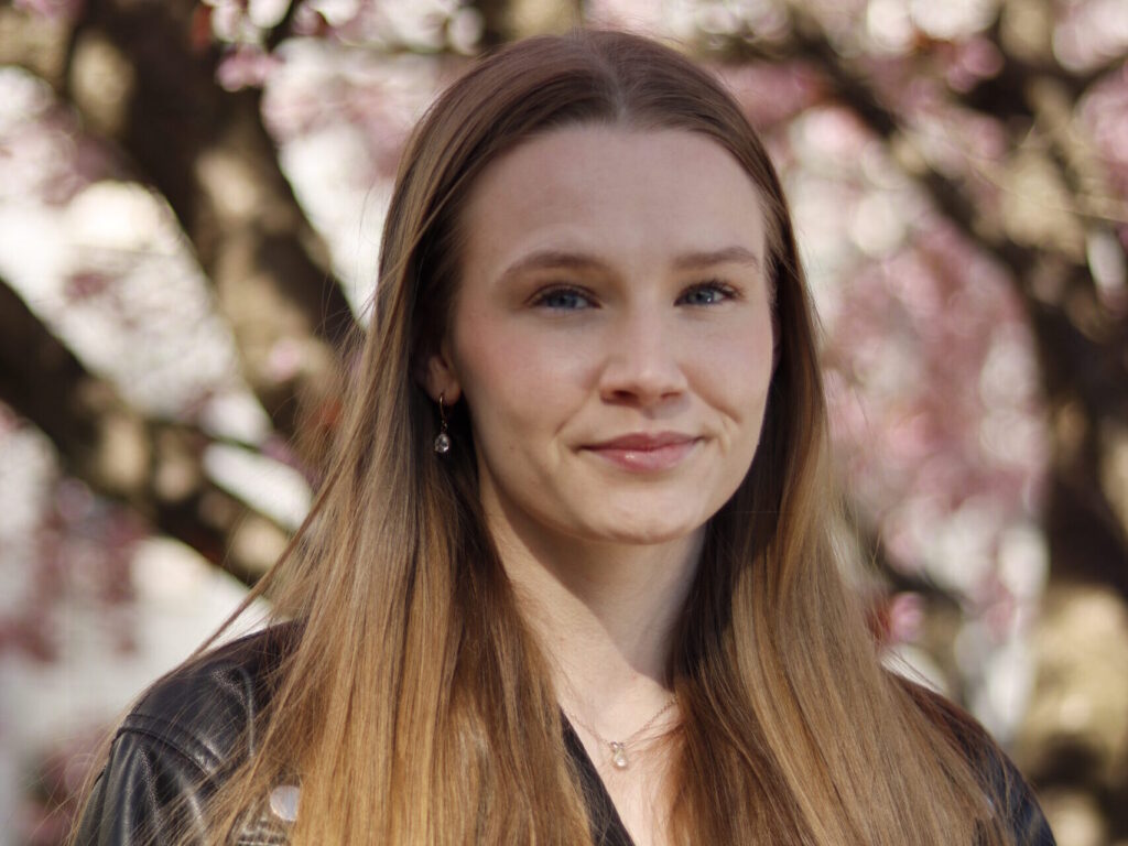 Jasmine vaaleat pitkät hiukset ja nahkatakki hymyilee kirsikkapuut taustalla.