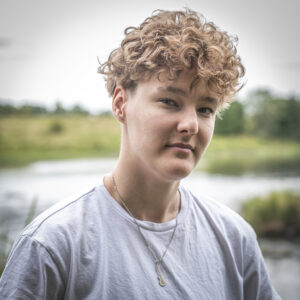 Jayden hymyilee joenvarsimaisemassa kesällä, kiharat hiukset ja valkoinen paita.