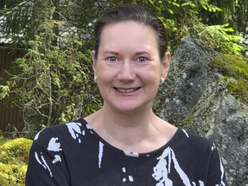 Päivi hymyilee ja katsoo kameraan kesäisessä puistossa. 