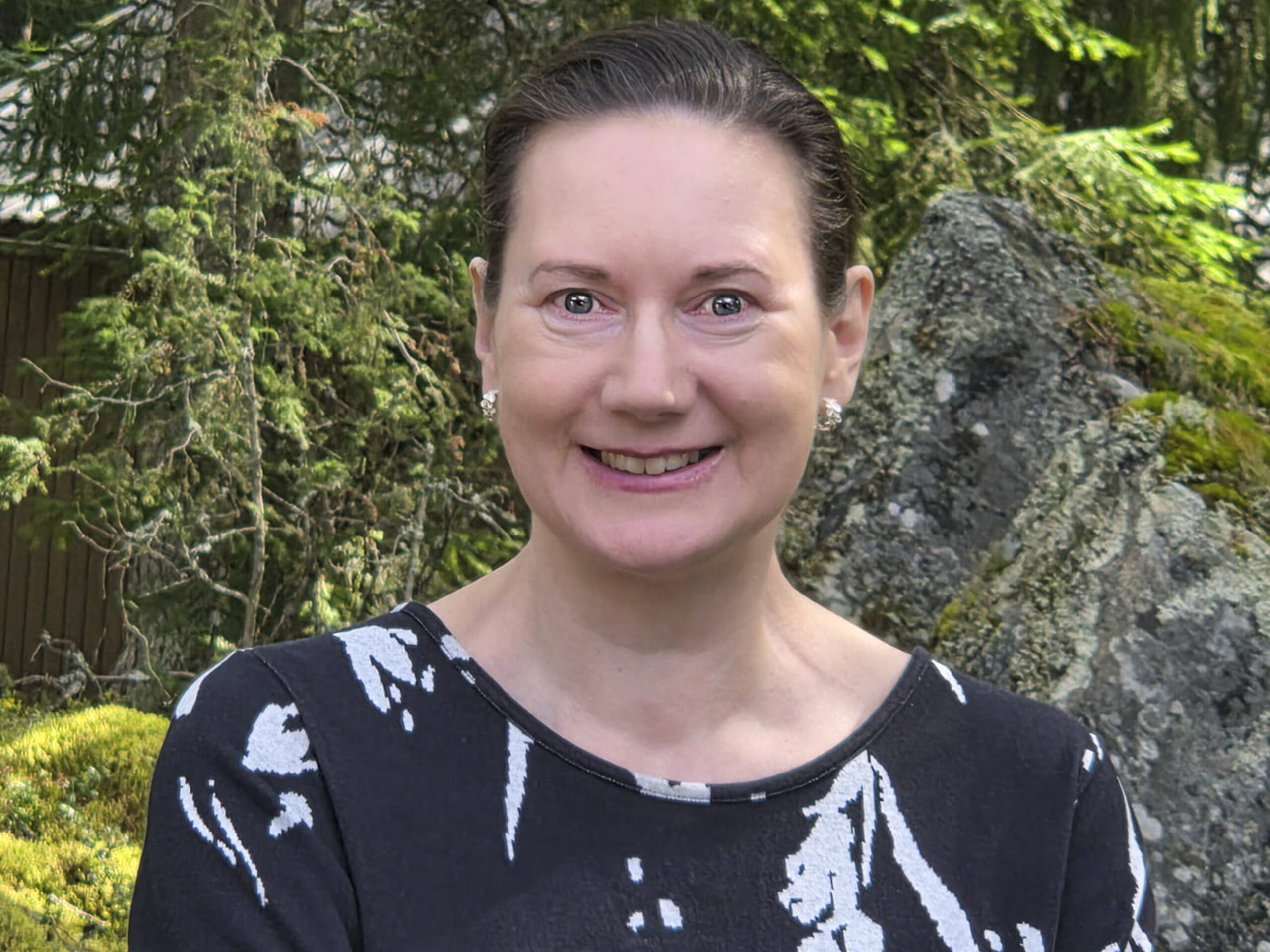 Päivi hymyilee ja katsoo kameraan kesäisessä puistossa.